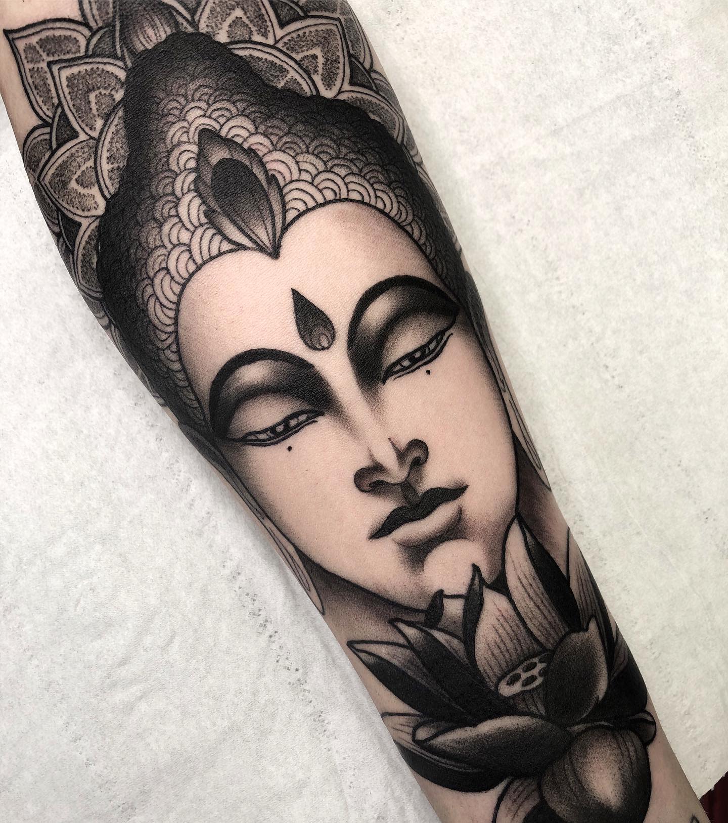 Black and gray tattoo of a serene Buddha face with a lotus flower on an arm